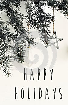 Happy Holidays text sign on stylish christmas branches with glass modern ornaments hanging on white wall. Creative christmas
