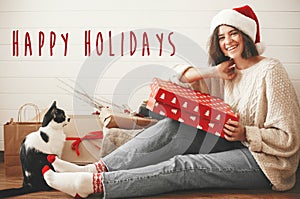 Happy Holidays text sign on stylish happy girl in santa hat and cozy sweater playing with cat and holding christmas gift box in
