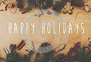 Happy Holidays text sign on stylish rustic frame of pine branches, cones, gingerbread cookies,thread, cinnamon, anise on rural