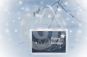 Happy holidays written on black chalk board hanging from a tree on blue, snowy background. Christmas tree ornament