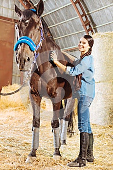 Happy horsewoman standing in big stable and cleaning horse