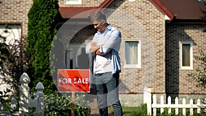 Happy house owner looking at for sale signboard against house, moving concept