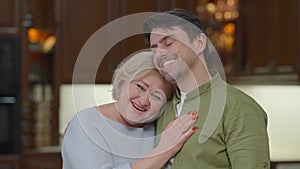 Happy hugging senior mother and adult son talking smiling standing at home indoors. Portrait of carefree Caucasian man