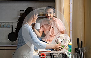 Happy husband having coffee while spending time with wife while busy washing washing vegetables for cooking at kitchen -