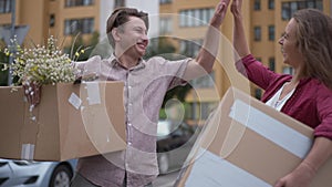 Happy husband and wife gesturing high-five in slow motion standing with parcels at building moving in new apartment