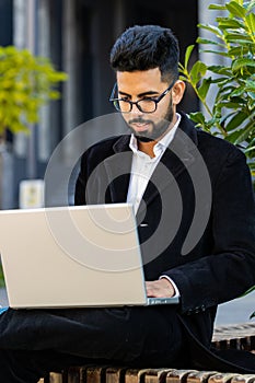 Happy Indian businessman typing working remote on wireless laptop computer browsing online outdoors