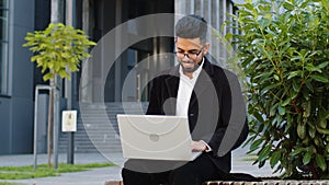 Happy Indian businessman typing working remote on wireless laptop computer browsing online outdoors