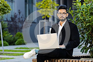 Happy Indian businessman typing working remote on wireless laptop computer browsing online outdoors