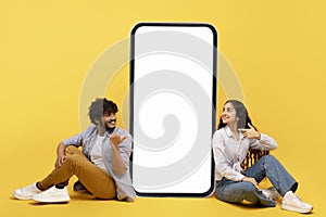 Happy indian couple sitting near big smartphone with blank white screen, demonstrating copy space for app or ad