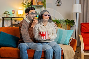 Happy Indian family couple in 3D glasses eating popcorn and watching comedy movie sitting on sofa