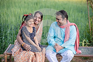 Happy Indian farmer family sitting on traditional bed at agricultural field in village, Young couple with their daughter in