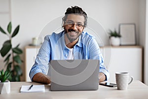 Happy Indian Freelancer Man Sitting At Desk With Laptop, Smiling At Camera