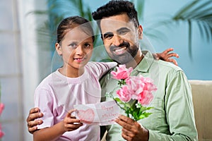 Happy indian girl kid giving gift with flowers to father by looking at camera at home - concept of fathers day, family