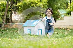 Happy indian girl kid painting cardboard toy house using brush at park - concept of childhood creativity, playful and