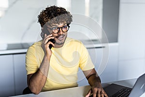 Happy indian guy having conversation on phone, using laptop at kitchen