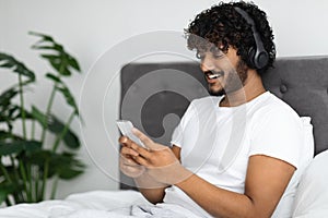 Happy indian guy sitting in bed with smartphone and headset