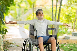 Happy indian kid with disability feeling fresh air by stretching hands sitting on wheelchair at park - concept of