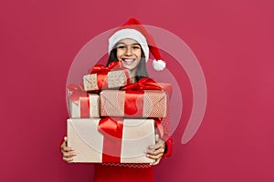 Happy indian kid girl wear santa hat holding many gifts boxes on red background.