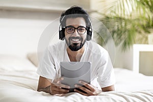 Happy Indian Man With Digital Tablet And Wireless Headphones Relaxing In Bed