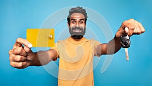 Happy indian man showing new car key and credit card, buying automobile, standing over blue studio background, panorama