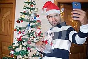 Happy Indian Man Wearing Santa hat Talking Selfie With Smart Phone Holding Christmas Gift Boxes Or Presents Enjoys Holiday Season
