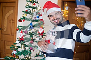 Happy Indian Man Wearing Santa hat Talking Selfie Or Video Call Over Smart Phone Holding Christmas Gift Boxes Or Presents Enjoys