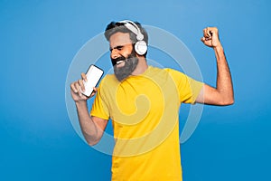 Happy indian man in wireless headphones singing at smartphone with empty screen in imaginary microphone, blue background