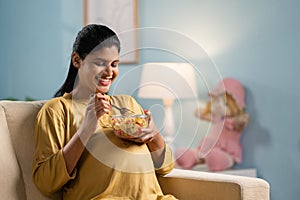 Happy indian pregnant woman eating fruits salad while sitting on sofa at home - concept of healthy lifestyles, Maternal