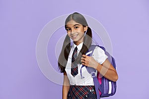 Happy indian school girl wearing uniform holding backpack on lilac background.