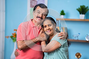 Happy Indian senior couple closely standing by embracing each other by looking camera at home - concept of family