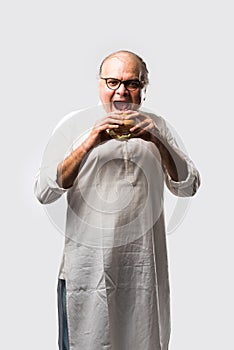 Happy Indian senior man eating burger or hamburger with cold drink