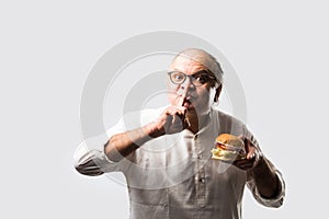 Happy Indian senior man eating burger or hamburger with cold drink