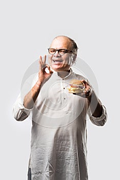 Happy Indian senior man eating burger or hamburger with cold drink