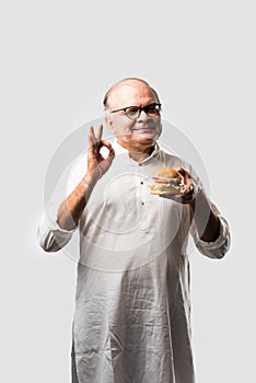 Happy Indian senior man eating burger or hamburger with cold drink