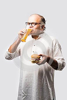 Happy Indian senior man eating burger or hamburger with cold drink