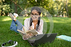 Happy indian student girl in eyeglasses reading book outdoors, preparing for exams and enjoying studying in park