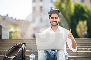 Happy indian student sitting on the stairs showing thumb up working on laptop, in the university campus. Technology, education and