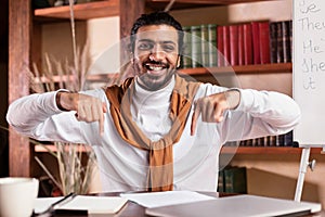 Happy Indian Teacher In Headphones Pointing At His Desk Indoors