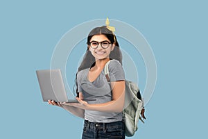 Happy Indian teen girl with backpack and laptop computer participating in online lesson on blue studio background