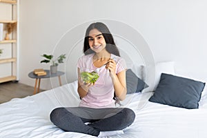 Happy Indian woman sitting on bed and eating tasty vegetable salad at home. Healthy lifestyle