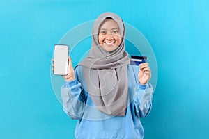 Happy Indonesian muslim woman showing a smart phone screen and holding debit card, isolated over blue background