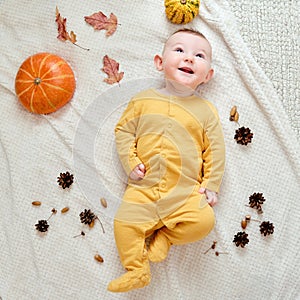 Happy infant baby lies on a blanket in yellow autumn clothes with a red pumpkin. Smiling child with orange leaves, top view