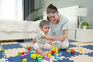 happy infant baby playing wooden block toy with mother on jigsaw mat in bedroom
