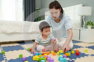 happy infant baby playing wooden block toy with mother on jigsaw mat in bedroom
