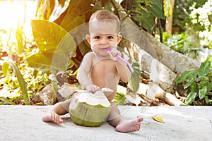 Happy infant baby at summer tropical vacation. Eats and drinks green young coconut. Sits on a ground and hold drinking straw and