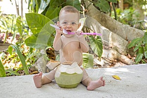 Happy infant baby is teething and gnaw a spoon. Eats and drinks green young coconut. Sits in the jungles