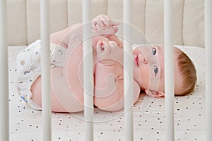 A happy infant baby with teething teeth in a diaper pulls his hand into his mouth. Funny child in a crib