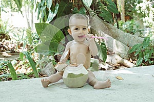 Happy infant baby at tropical vacation. Eats and drinks green young coconut. Sits on a ground and hold spoon and drinking straw.