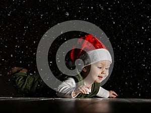 Happy infant boy toddler in red Santa hat and white - green clothes is lying on floor looking at copy space under snow