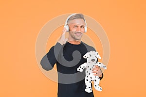 Happy infantile middle-aged man play with toy dog listening to music yellow background, headphones